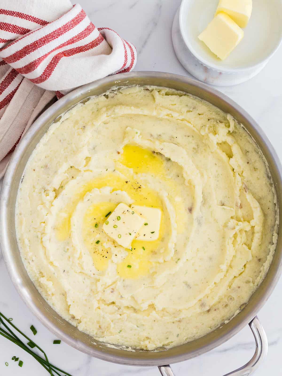 A pot of mashed potatoes topped with butter and chives, next to a red striped towel.