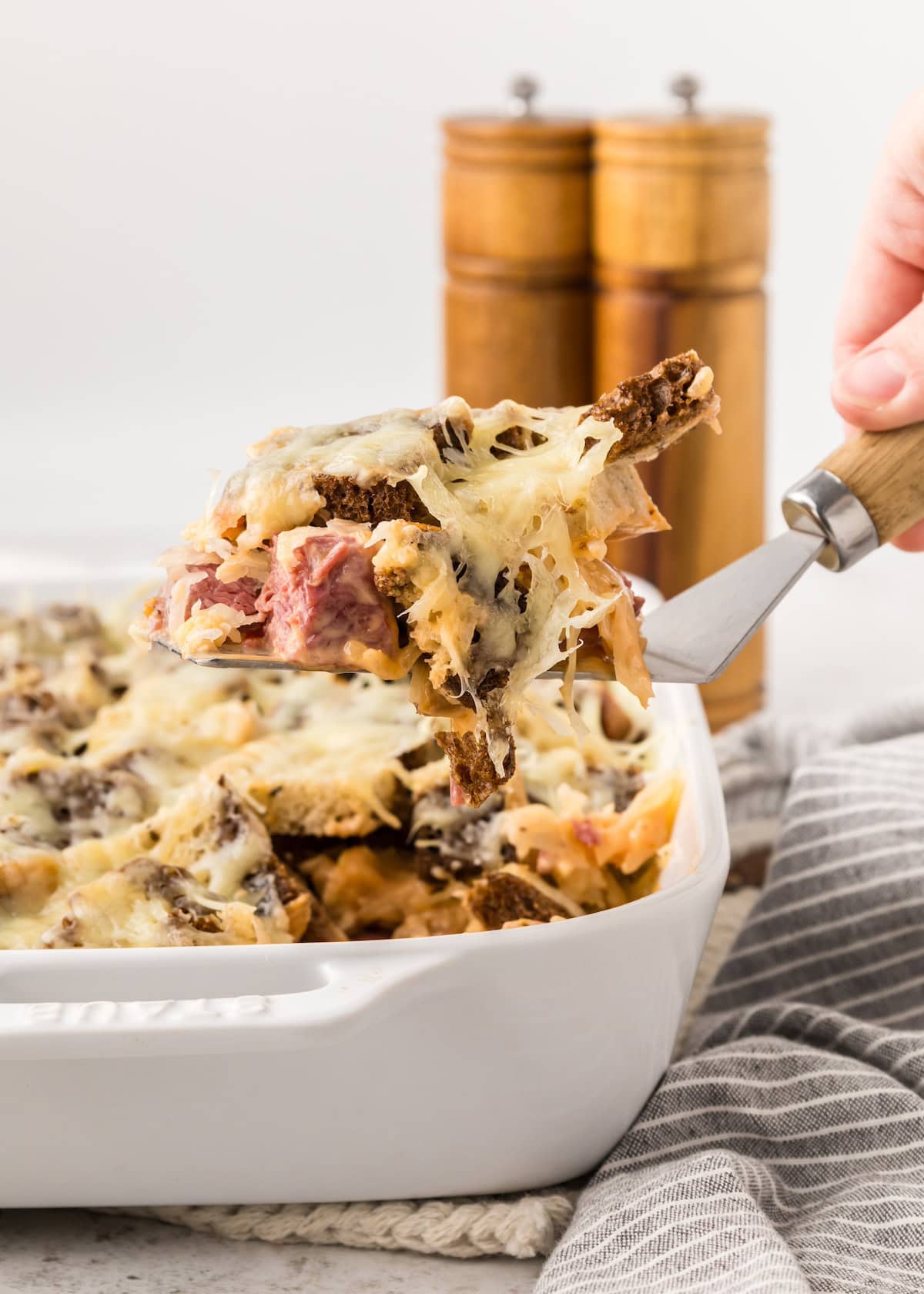 A hand serves cheesy baked casserole from a white dish with shredded cheese on top.