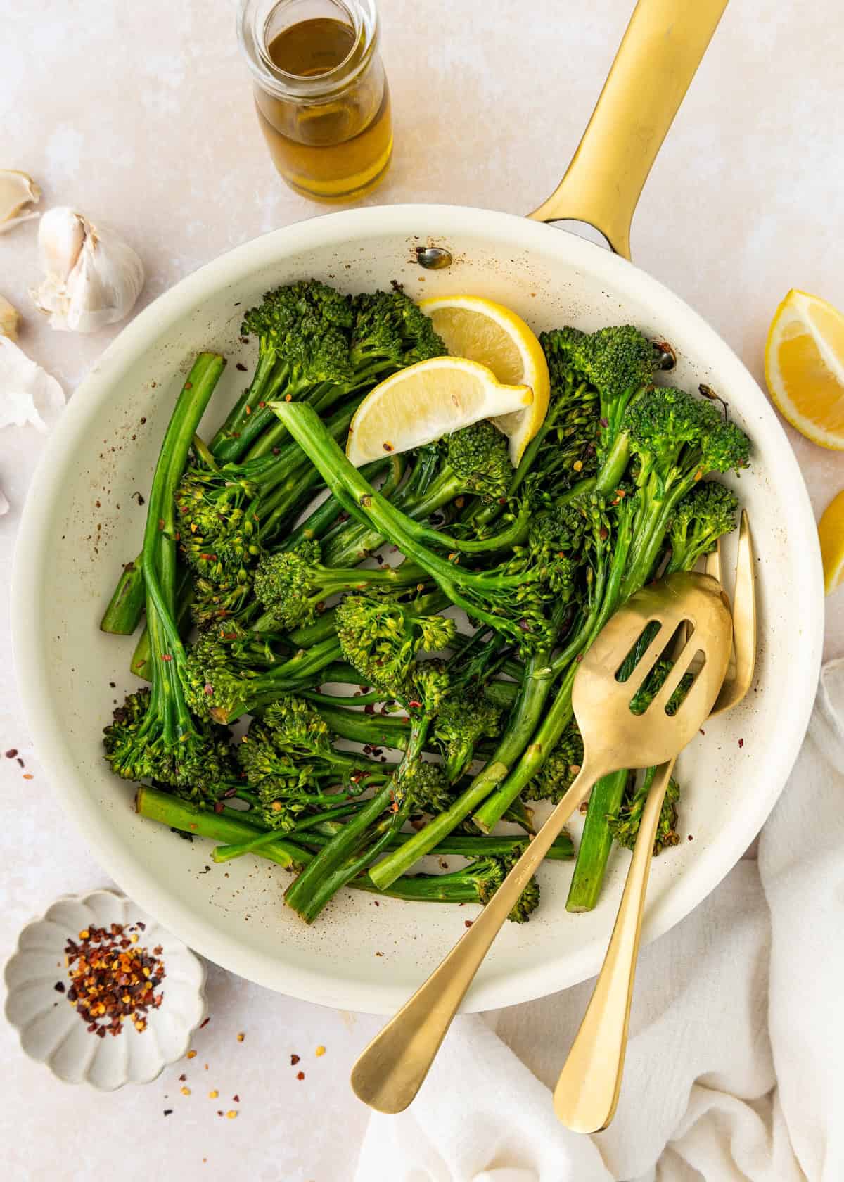 Cooked broccolini in a skillet with lemon wedges and gold serving utensils.