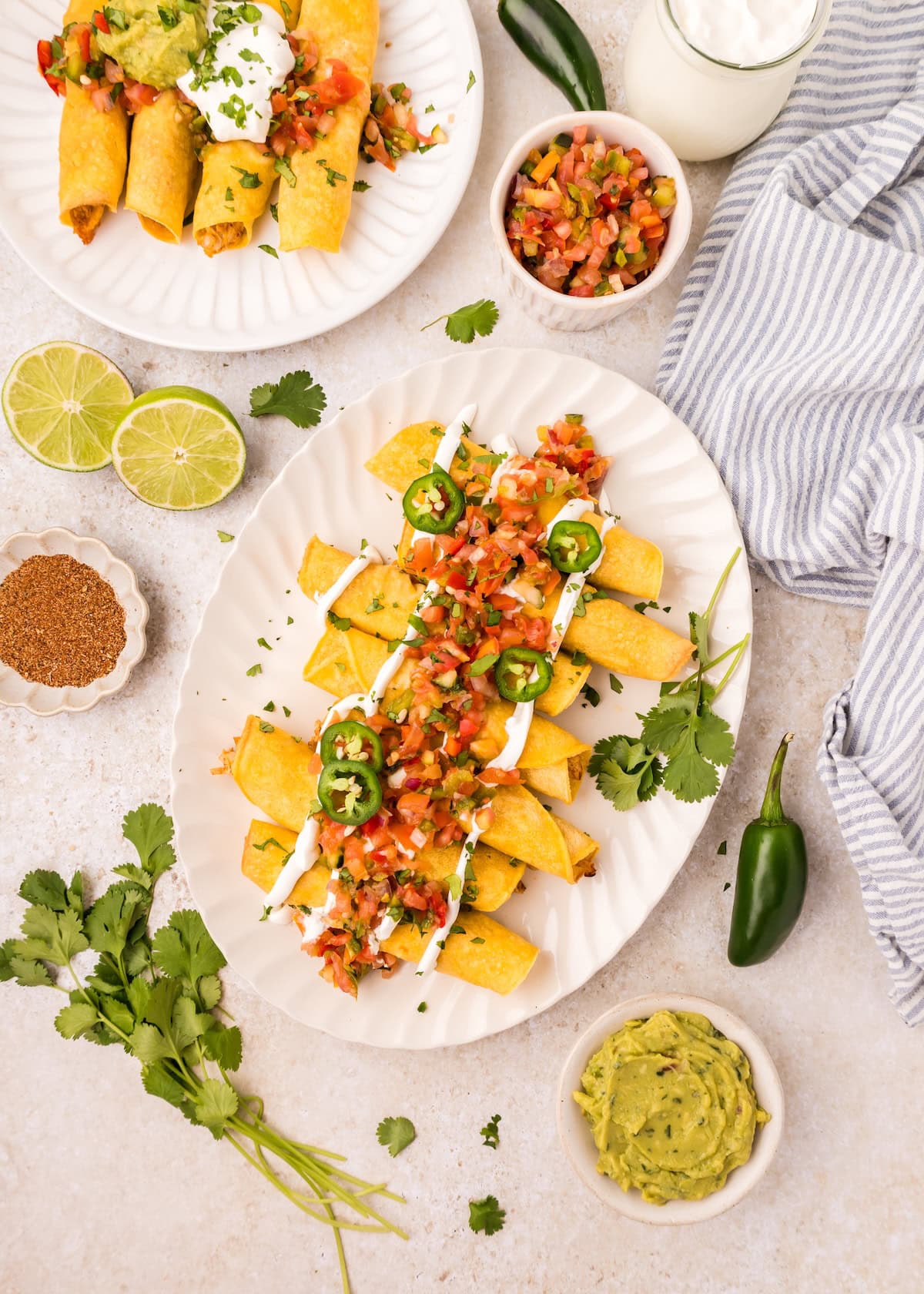 Plates of taquitos topped with salsa, jalapeños, and sour cream, with sides of guacamole and limes.