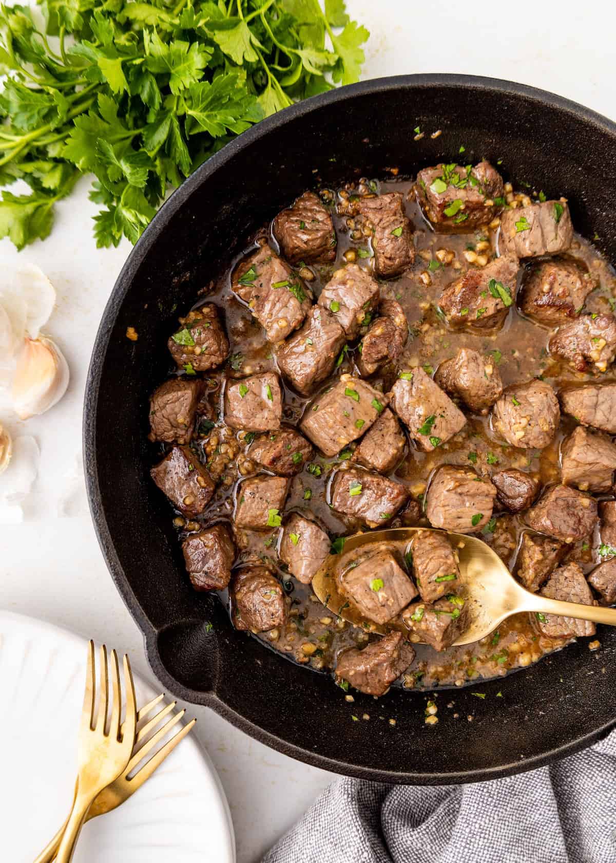 Cubes of cooked steak in a skillet with herbs, next to parsley and garlic on a white surface.