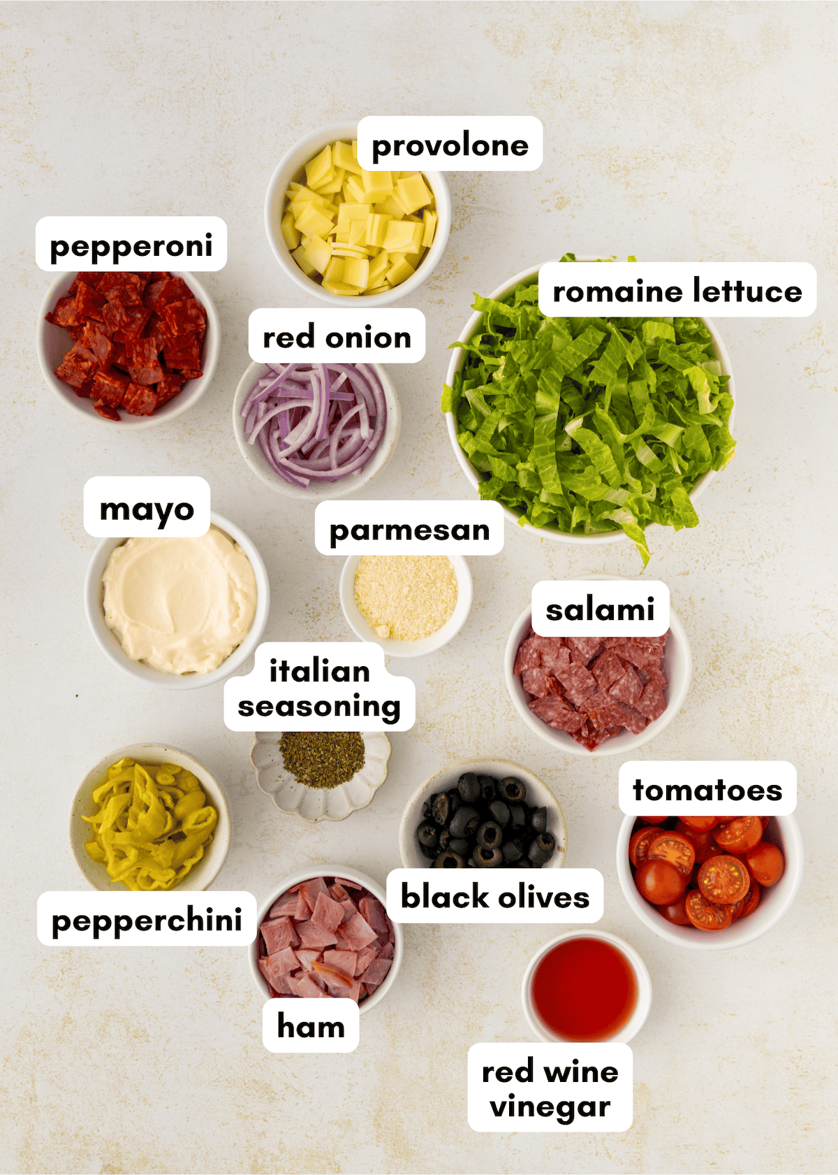 Ingredients for an Italian sub salad arranged in bowls and labeled on a light surface.