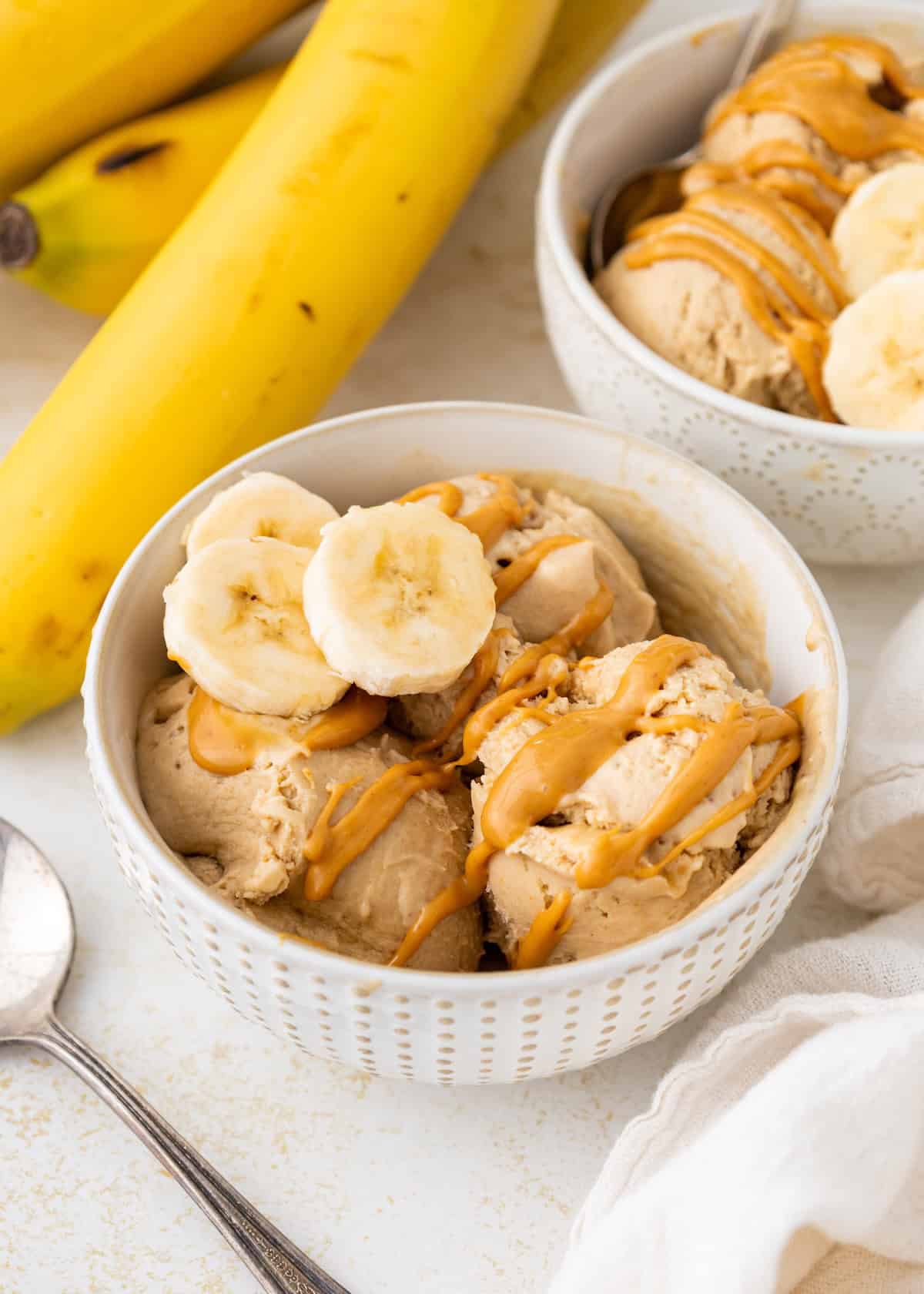 Bowl of banana ice cream topped with banana slices and peanut butter drizzle, with bananas nearby.