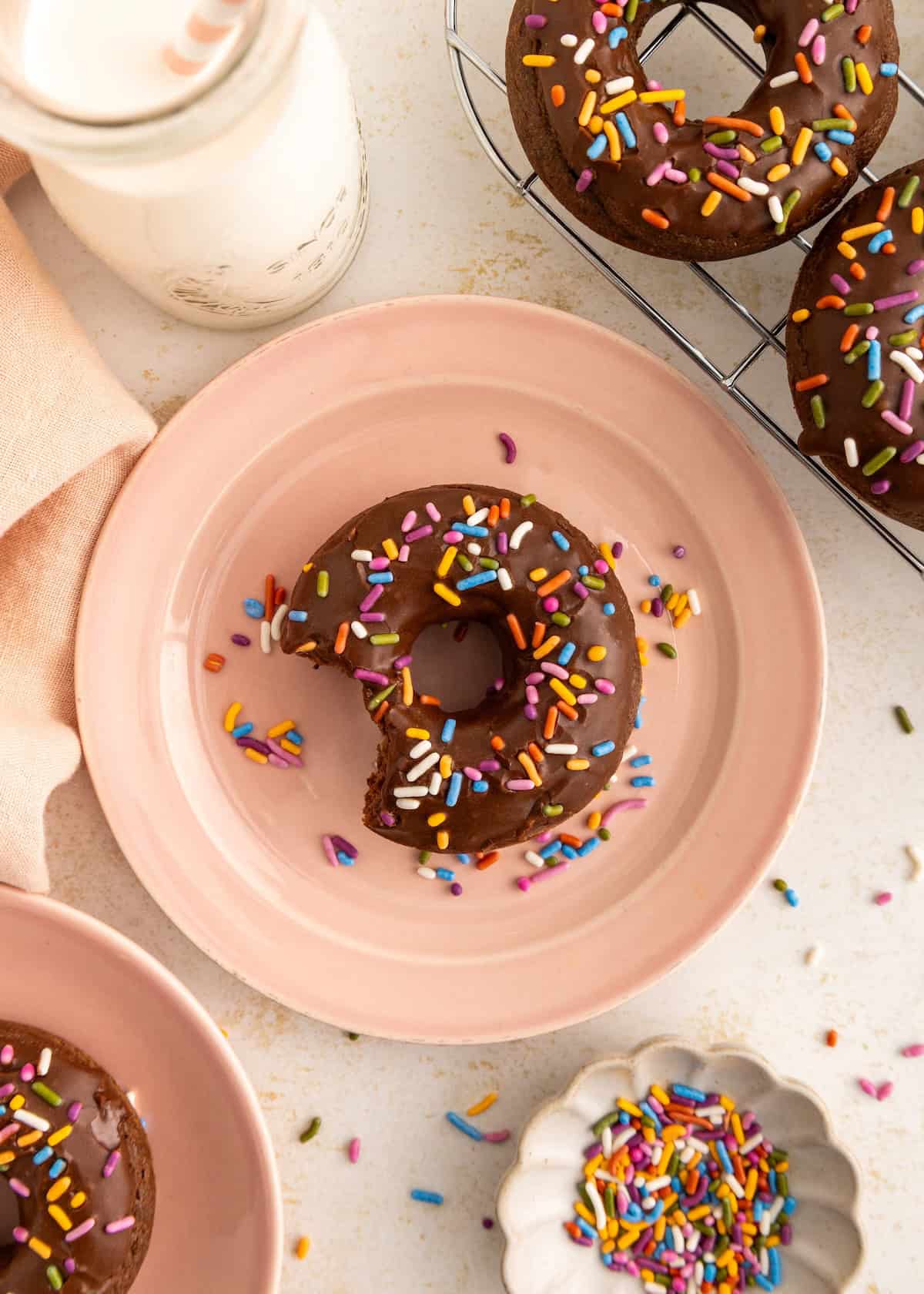 A chocolate donut with sprinkles and a bite taken out, on a pink plate beside a bottle of milk.