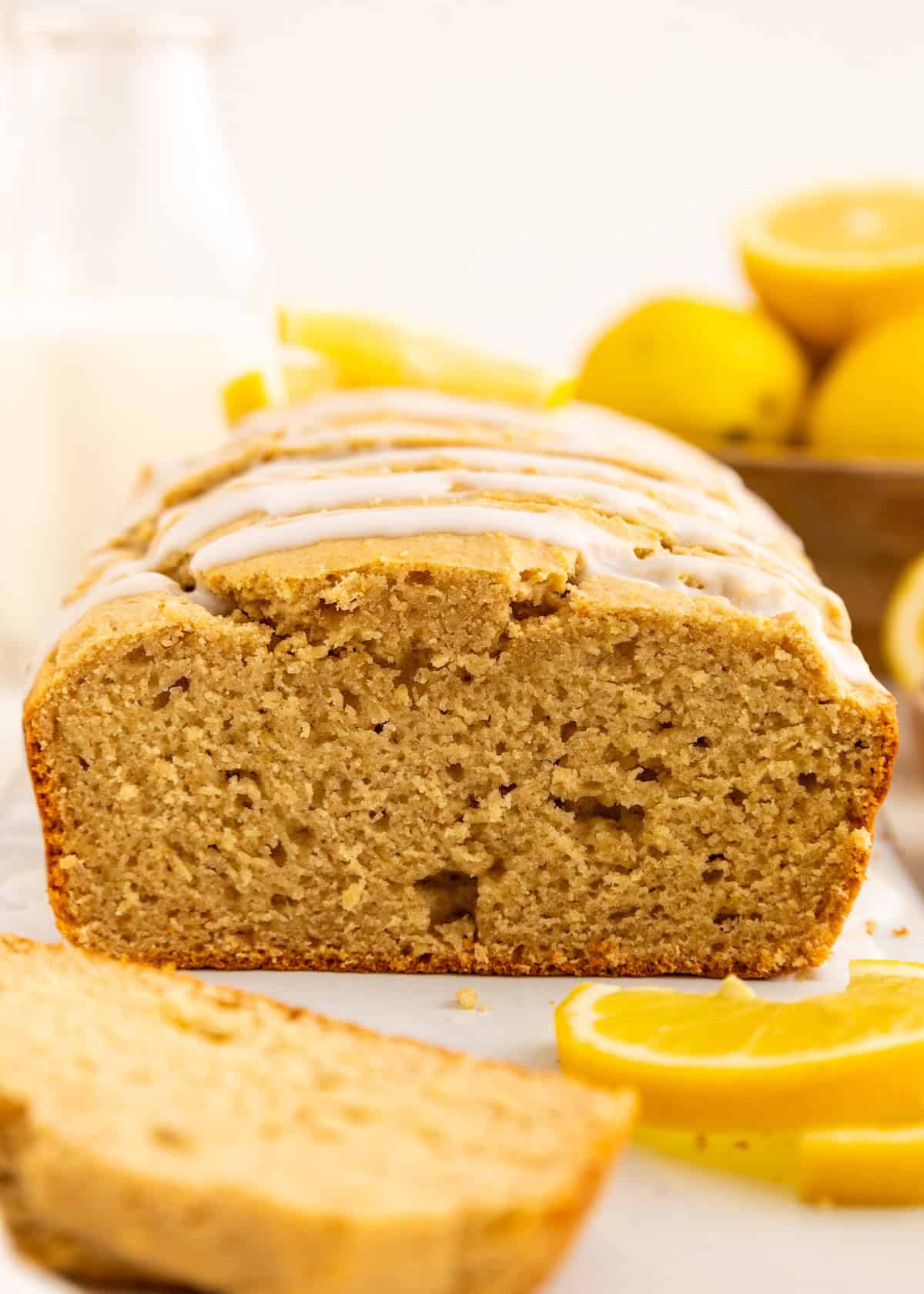 A sliced loaf of glazed lemon bread with lemons and a bottle of milk in the background.