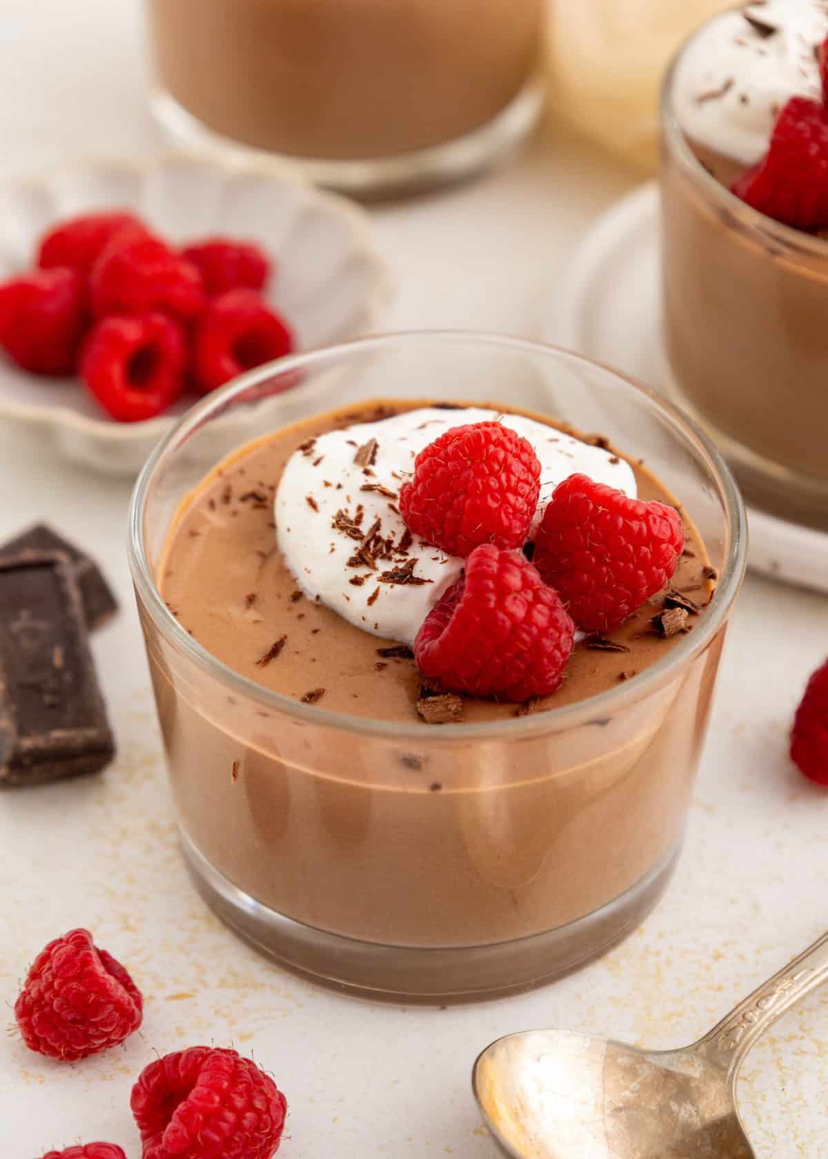 A glass of chocolate mousse topped with whipped cream, raspberries, and chocolate shavings.
