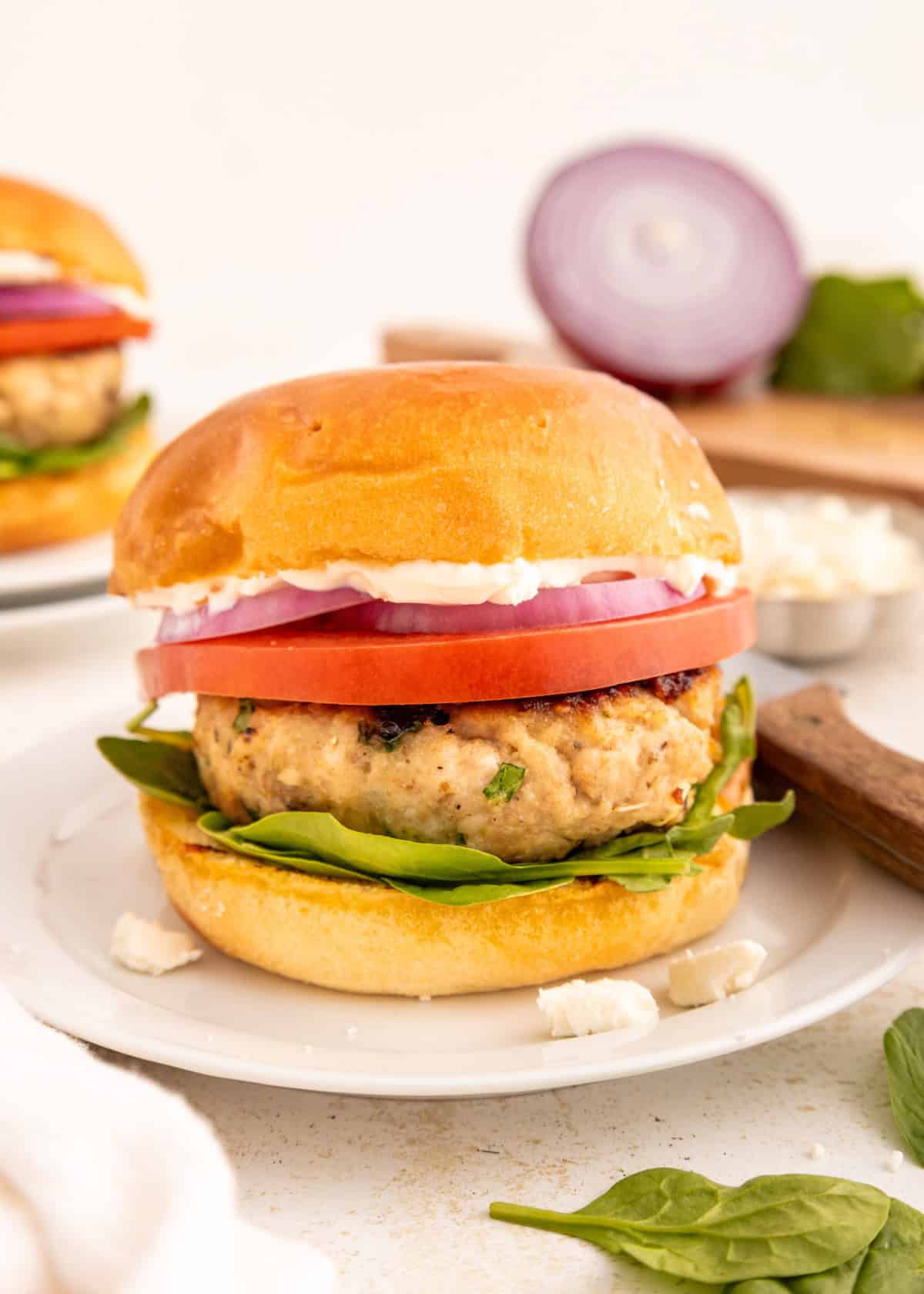A chicken burger with spinach, tomato, red onion, and feta on a white plate.
