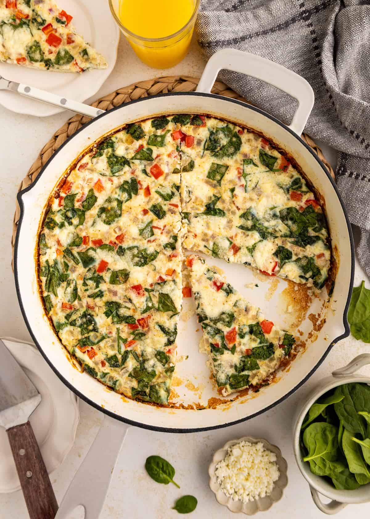 A vegetable frittata in a skillet with one slice removed, surrounded by fresh ingredients.