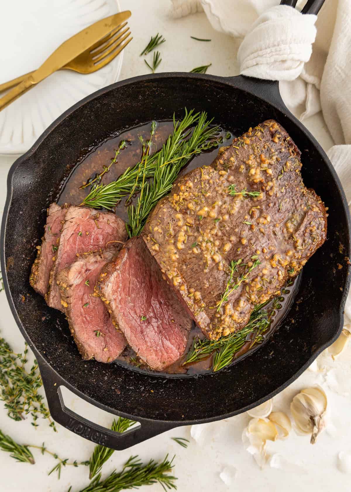 Garlic herb roast beef sliced in a skillet with herbs and garlic, ready to serve with plates.