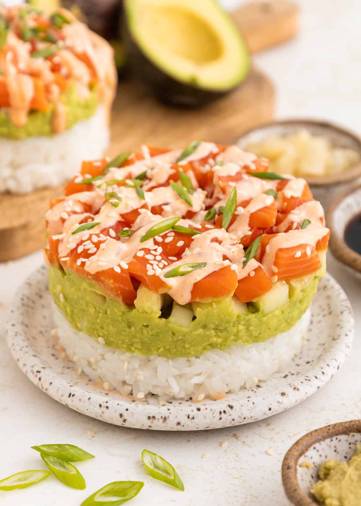 Stacked sushi stacks with rice, avocado, and salmon, topped with sauce, sesame seeds, and green onions.