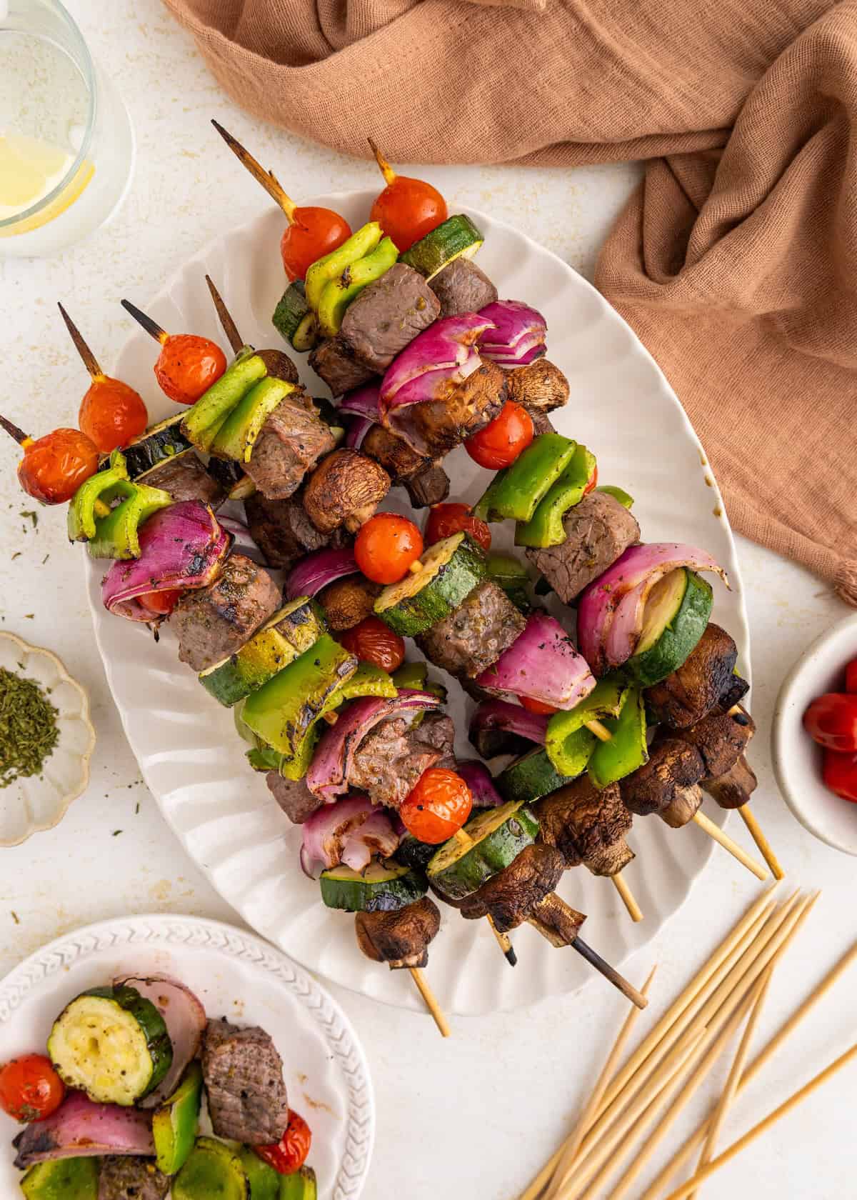 Grilled beef, bell pepper, tomato, onion, and mushroom skewers on a white platter.